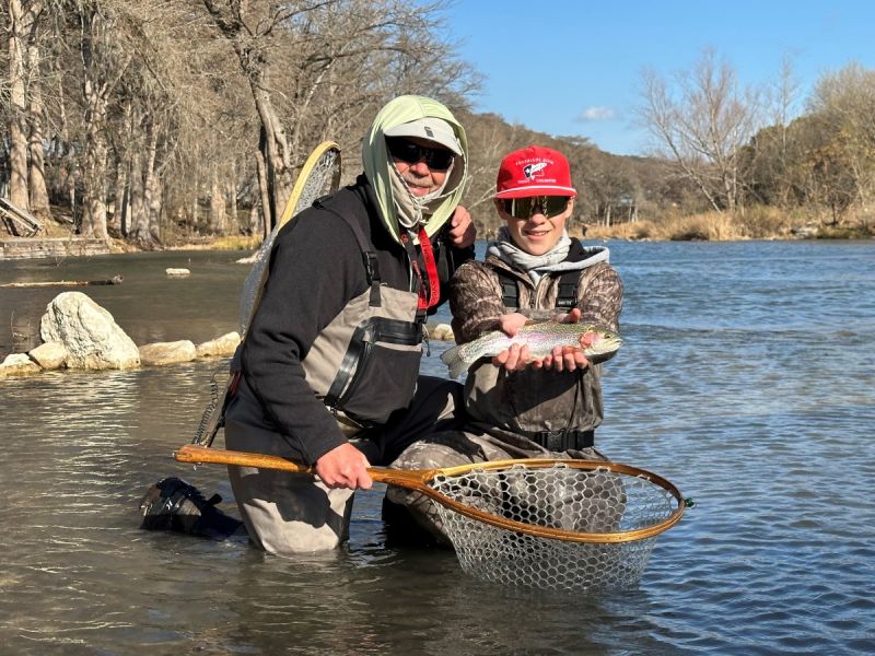 A young angler and a mentor kneeling in a Hill Country river with a netted rainbow trout